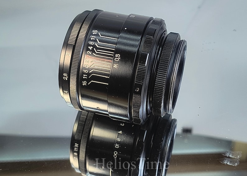 Puede incluir: Un objetivo de c&aacute;mara negro con marcas blancas y un anillo de enfoque estriado. El objetivo tiene una serie de n&uacute;meros y l&iacute;neas que indican los ajustes de apertura. El objetivo se refleja en una superficie espejada.