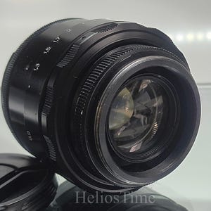 Puede incluir: Un objetivo de c&aacute;mara negro con un anillo de enfoque estriado y marcas de apertura. El objetivo tiene un elemento de vidrio transparente y se ve una tapa de objetivo. El texto "Helios Time" est&aacute; en la parte inferior.