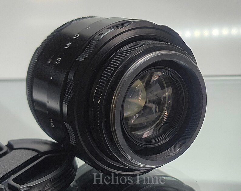 Puede incluir: Un objetivo de c&aacute;mara negro con un anillo de enfoque estriado y marcas de apertura. El objetivo tiene un elemento de vidrio transparente y se ve una tapa de objetivo. El texto "Helios Time" est&aacute; en la parte inferior.