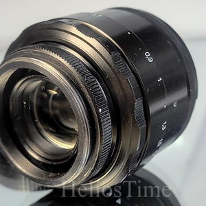 Puede incluir: Un objetivo de c&aacute;mara negro con anillos y marcas plateadas. El objetivo tiene un anillo de enfoque estriado y una escala con n&uacute;meros. El objetivo est&aacute; en &aacute;ngulo, mostrando sus elementos internos. Producto para fotograf&iacute;a.