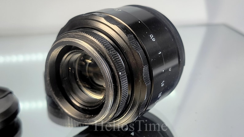 Puede incluir: Un objetivo de c&aacute;mara negro con anillos y marcas plateadas. El objetivo tiene un anillo de enfoque estriado y una escala con n&uacute;meros. El objetivo est&aacute; en &aacute;ngulo, mostrando sus elementos internos. Producto para fotograf&iacute;a.