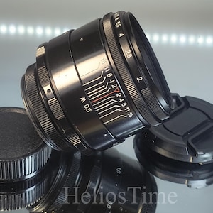 Puede incluir: Lente de c&aacute;mara negra con una tapa de lente y una tapa protectora. La lente tiene marcas blancas y rojas para la configuraci&oacute;n de la apertura y un anillo de enfoque. La lente est&aacute; sobre una superficie reflectante.