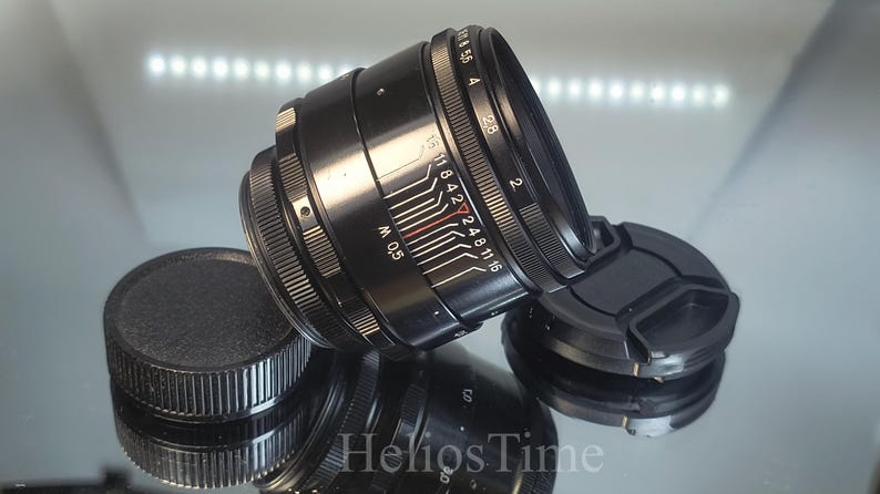 Puede incluir: Lente de c&aacute;mara negra con una tapa de lente y una tapa protectora. La lente tiene marcas blancas y rojas para la configuraci&oacute;n de la apertura y un anillo de enfoque. La lente est&aacute; sobre una superficie reflectante.