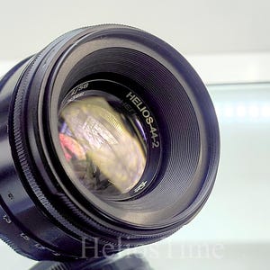 Puede incluir: Un objetivo de c&aacute;mara Helios-44-2 negro con un elemento de vidrio reflectante. El objetivo tiene un anillo de enfoque estriado y marcas de apertura y distancia. El texto "Helios-44-2" es visible.
