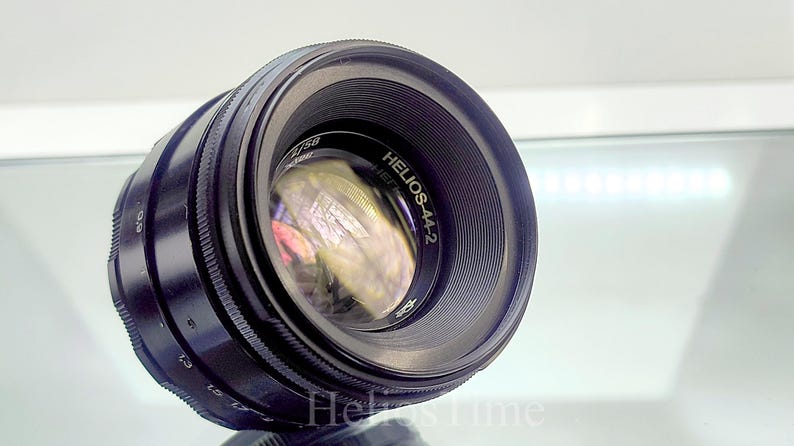 Puede incluir: Un objetivo de c&aacute;mara Helios-44-2 negro con un elemento de vidrio reflectante. El objetivo tiene un anillo de enfoque estriado y marcas de apertura y distancia. El texto "Helios-44-2" es visible.