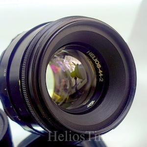 Puede incluir: Un objetivo de c&aacute;mara negro Helios-44-2 con interior reflectante. El objetivo tiene un anillo de enfoque estriado y el texto "Helios-44-2" en letras doradas. Una tapa de objetivo negra es visible en primer plano.