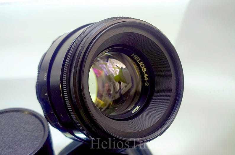 Puede incluir: Un objetivo de c&aacute;mara negro Helios-44-2 con interior reflectante. El objetivo tiene un anillo de enfoque estriado y el texto "Helios-44-2" en letras doradas. Una tapa de objetivo negra es visible en primer plano.