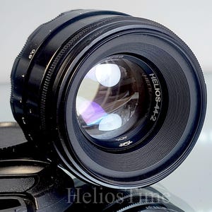 Puede incluir: Un objetivo de c&aacute;mara negro Helios-44-2 con un elemento de vidrio reflectante. El objetivo tiene un anillo de enfoque estriado y el texto "Helios-44-2" es visible. Una tapa de objetivo negra est&aacute; en primer plano.
