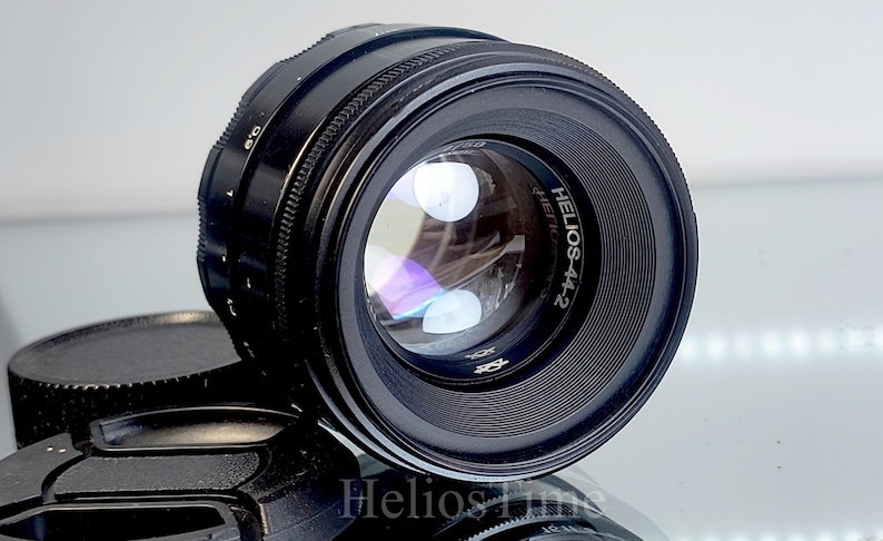 Puede incluir: Un objetivo de c&aacute;mara negro Helios-44-2 con un elemento de vidrio reflectante. El objetivo tiene un anillo de enfoque estriado y el texto "Helios-44-2" es visible. Una tapa de objetivo negra est&aacute; en primer plano.