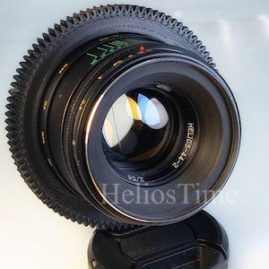 May include: A black Helios-44-2 camera lens with a ridged focusing ring. The lens has green and red markings, with the text "Helios-44-2" and "HeliosTime" visible. The lens is mounted on a black base.
