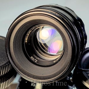 Puede incluir: Primer plano de un objetivo negro Helios-44-2 con un elemento de vidrio reflectante. El objetivo tiene un anillo de enfoque estriado y el texto "HELIOS-44-2 2/58" es visible. Una tapa de objetivo es parcialmente visible.