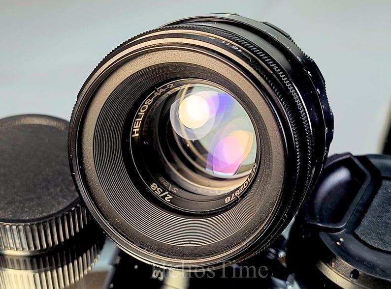 Puede incluir: Primer plano de un objetivo negro Helios-44-2 con un elemento de vidrio reflectante. El objetivo tiene un anillo de enfoque estriado y el texto "HELIOS-44-2 2/58" es visible. Una tapa de objetivo es parcialmente visible.