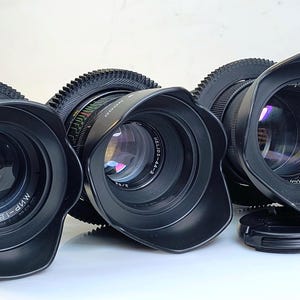 May include: Three black camera lenses with attached gear rings and lens hoods. The lenses are arranged in a row, with the front lens displaying the text "MИP-1B 2.8/37". The lenses are designed for professional photography.