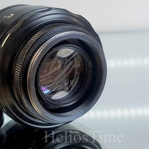 Puede incluir: Un objetivo de c&aacute;mara negro con marcas plateadas y un anillo de enfoque estriado. El objetivo tiene un elemento de vidrio transparente y est&aacute; parcialmente cubierto por una tapa de objetivo negra. El texto "Helios Time" es visible.