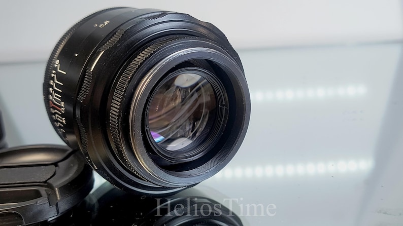 Puede incluir: Un objetivo de c&aacute;mara negro con marcas plateadas y un anillo de enfoque estriado. El objetivo tiene un elemento de vidrio transparente y est&aacute; parcialmente cubierto por una tapa de objetivo negra. El texto "Helios Time" es visible.
