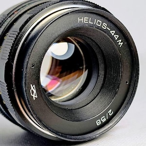 May include: A black camera lens with a textured focus ring and a metal front element. The front rim is clearly marked with the white text HELIOS-44M and 2/58, along with a small geometric logo. The glass element reflects light internally.
