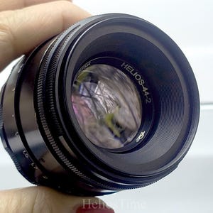 Puede incluir: Un objetivo de c&aacute;mara negro Helios-44-2 con un anillo de enfoque estriado. La lente tiene el texto "Helios-44-2" y "58" impreso. La lente se sostiene en una mano.