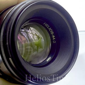 Puede incluir: Un objetivo de c&aacute;mara negro Helios-44-2 con un elemento de vidrio reflectante. El objetivo tiene un anillo de enfoque estriado y el texto "Helios-44-2" es visible en el cuerpo del objetivo. El objetivo se sostiene sobre un fondo blanco.