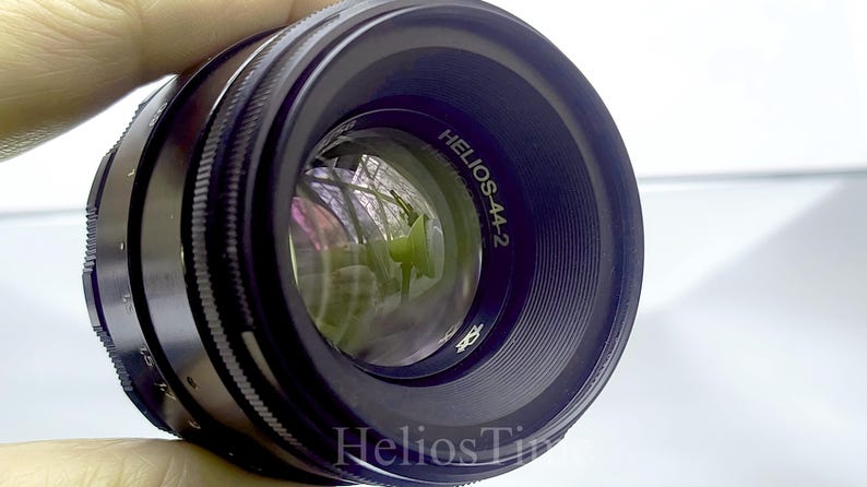 Puede incluir: Un objetivo de c&aacute;mara negro Helios-44-2 con un elemento de vidrio reflectante. El objetivo tiene un anillo de enfoque estriado y el texto "Helios-44-2" es visible en el cuerpo del objetivo. El objetivo se sostiene sobre un fondo blanco.