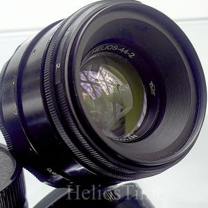 Puede incluir: Un objetivo de c&aacute;mara negro Helios-44-2 con una tapa de objetivo. El objetivo tiene una superficie reflectante con el texto "Helios-44-2" y otras marcas. La tapa del objetivo es negra y texturizada.
