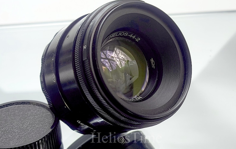Puede incluir: Un objetivo de c&aacute;mara negro Helios-44-2 con una tapa de objetivo. El objetivo tiene una superficie reflectante con el texto "Helios-44-2" y otras marcas. La tapa del objetivo es negra y texturizada.