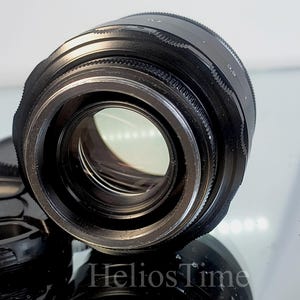 Puede incluir: Un objetivo de c&aacute;mara negro con un elemento de vidrio transparente, acompa&ntilde;ado de una tapa de objetivo. El objetivo tiene un anillo de enfoque estriado y est&aacute; sobre una superficie reflectante. La imagen est&aacute; bien iluminada.