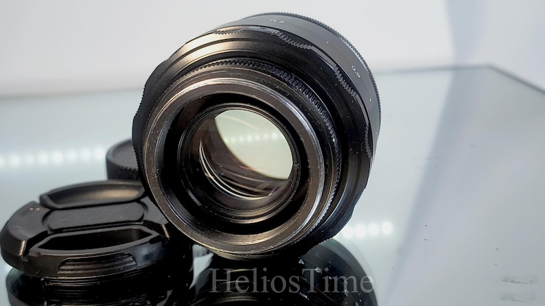 Puede incluir: Un objetivo de c&aacute;mara negro con un elemento de vidrio transparente, acompa&ntilde;ado de una tapa de objetivo. El objetivo tiene un anillo de enfoque estriado y est&aacute; sobre una superficie reflectante. La imagen est&aacute; bien iluminada.