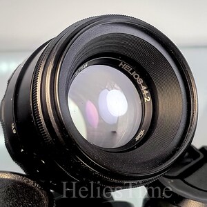 Puede incluir: Un objetivo de c&aacute;mara negro Helios-44-2 con un elemento de vidrio reflectante. El objetivo tiene un anillo de enfoque estriado y el texto "Helios-44-2" es visible. Una tapa de objetivo negra est&aacute; en primer plano.