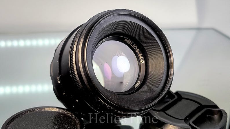 Puede incluir: Un objetivo de c&aacute;mara negro Helios-44-2 con un elemento de vidrio reflectante. El objetivo tiene un anillo de enfoque estriado y el texto "Helios-44-2" es visible. Una tapa de objetivo negra est&aacute; en primer plano.