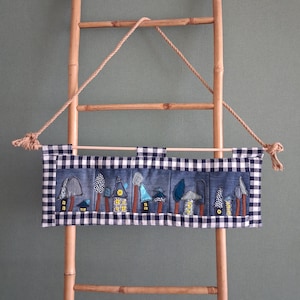 Peut inclure: Une tenture murale rectangulaire en tissu avec un panneau en jean bleu orné de motifs de maisons et d'arbres brodés et appliqués. L'ensemble est bordé d'un tissu à carreaux vichy noir et blanc et suspendu à une tige en bois par une corde.