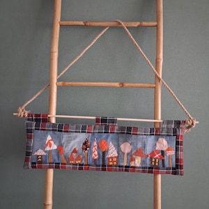 Peut inclure: Une tenture murale rectangulaire en tissu présentant une scène en patchwork de petites maisons et d'arbres aux motifs et couleurs variés, encadrée par une bordure à carreaux rouges, noirs et blancs. Suspendue à une tige en bois avec une corde beige.