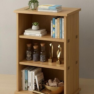 May include: A light oak bookshelf with three shelves, displaying books, decorative items, and small potted plants. The shelves hold glass jars with cork stoppers, gold sculptures, and a brass bowl.