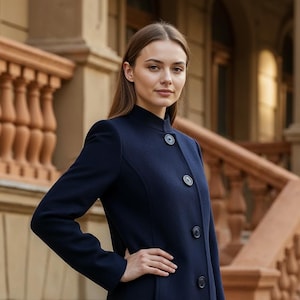 May include: A navy blue coat with a high collar and three dark buttons. The long coat has a tailored fit and a classic design. The model is wearing the coat in front of a building with a stone facade.