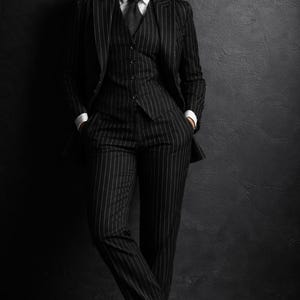 Puede incluir: Un traje a rayas finas negro con camisa blanca y corbata. El traje incluye blazer, chaleco y pantal&oacute;n. La persona lleva botas negras. El fondo es una pared gris oscuro.