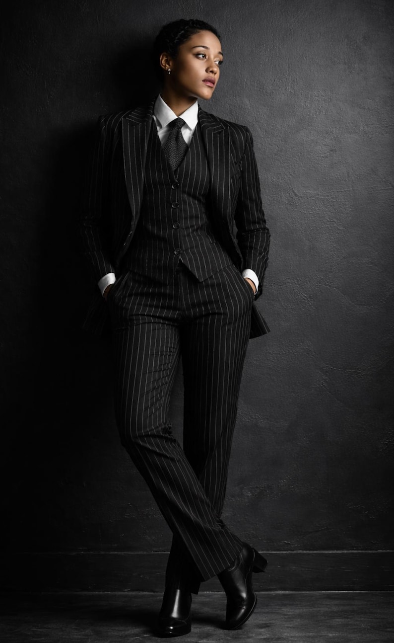Puede incluir: Un traje a rayas finas negro con camisa blanca y corbata. El traje incluye blazer, chaleco y pantal&oacute;n. La persona lleva botas negras. El fondo es una pared gris oscuro.