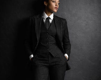May include: A black pinstripe suit, comprising a blazer, waistcoat, and trousers, paired with a white shirt and patterned tie. The person is leaning against a dark grey wall.