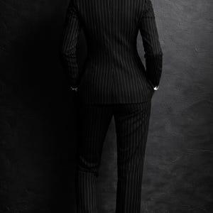 Puede incluir: Un traje negro de raya diplom&aacute;tica, compuesto por una americana y un pantal&oacute;n, se muestra desde atr&aacute;s. El traje tiene un dise&ntilde;o cl&aacute;sico con finas rayas blancas. Se ve una camisa blanca con cuello. El fondo es una pared oscura y texturizada.