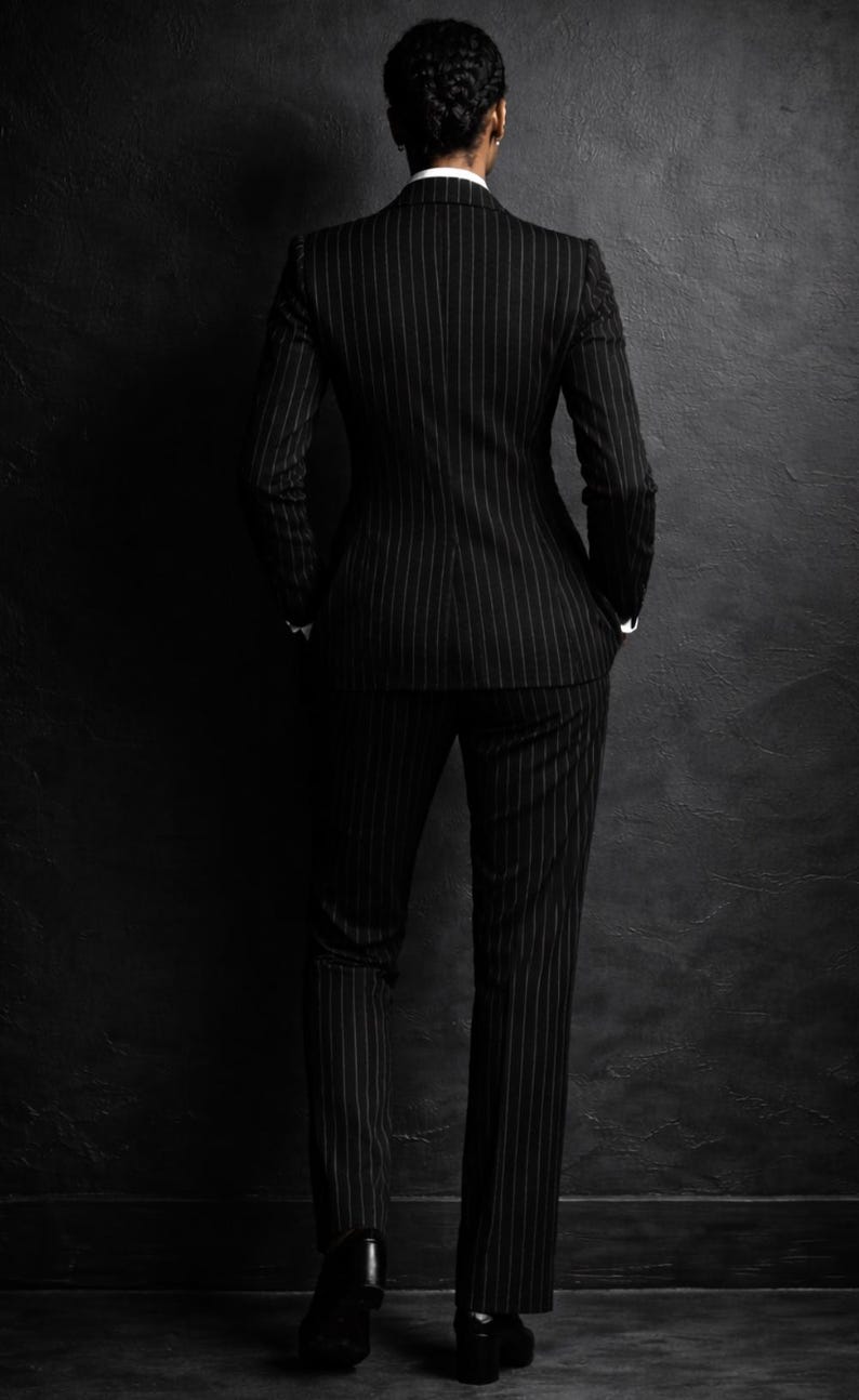 Puede incluir: Un traje negro de raya diplom&aacute;tica, compuesto por una americana y un pantal&oacute;n, se muestra desde atr&aacute;s. El traje tiene un dise&ntilde;o cl&aacute;sico con finas rayas blancas. Se ve una camisa blanca con cuello. El fondo es una pared oscura y texturizada.
