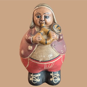 Puede incluir: Figura de arte popular pintada a mano de una persona tocando un instrumento de viento. La figura lleva un abrigo rojo, pantalones rojos y zapatos negros. El instrumento es de color beige con rayas negras. La figura tiene una cara beige y una cofia blanca.