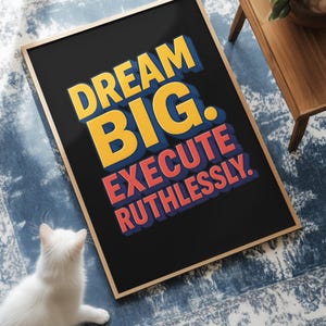 May include: A framed poster with the words "DREAM BIG. EXECUTE RUTHLESSLY." in yellow and red lettering on a black background. The poster is on a blue and white patterned rug. A white cat sits near the poster.