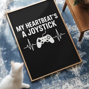 Puede incluir: Impresión artística enmarcada en negro con el texto blanco "MY HEARTBEAT'S A JOYSTICK", un gráfico de controlador blanco y una línea de latidos. Un gato blanco está cerca de la obra de arte sobre una alfombra azul estampada.