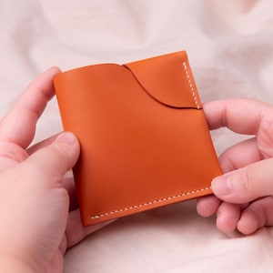 Könnte beinhalten: Ein kleines, orangefarbenes Lederportemonnaie mit geschwungenem Klappdeckel und weißen Nähten. Das Portemonnaie wird mit zwei Händen vor einem weichen, neutralen Hintergrund gehalten. Das Leder hat eine glatte Textur.