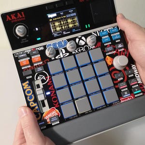 May include: A black and grey Akai MPC Sample music production device with a screen showing audio waveforms. Features logos for Sega, Xbox, EA, Konami, and Capcom. Includes buttons, knobs, and a speaker. The device is held by two hands.