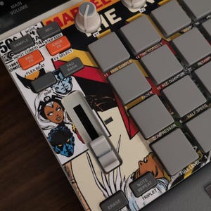 May include: Close-up of a Marvel Comics-themed music production device. The device has grey pads, knobs, and buttons with labels like "Sample" and "FX Select." Comic book art of Storm and other characters is visible.