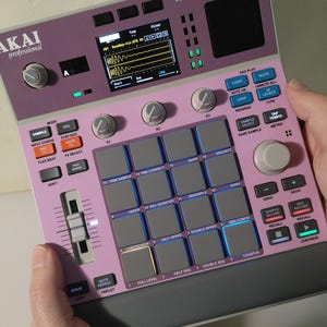 May include: A pink and grey Akai MPC Sample music production device. It features a screen, buttons, pads, and knobs for music creation. The device is held in a person's hands.