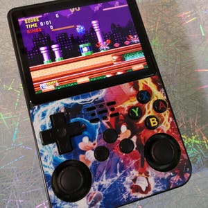 Może przedstawiać: Przenośna konsola do gier z grą Sonic the Hedgehog. Konsola ma czarną ramkę z kolorowymi przyciskami i grafiką Sonic. Ekran wyświetla wynik, czas i pierścienie gry.