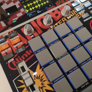 May include: A digital music sampler featuring a grid of sixteen grey square pads, three grey control knobs, and a vertical fader. The device has a black, red, and white graphic surface with the text Nintendo and Lofi visible on the casing.
