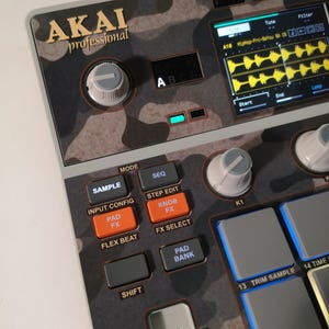 May include: An AKAI Professional music production controller with a camouflage pattern. The image highlights buttons like "SAMPLE" and "KNOB FX", and a screen showing audio waveforms. The design includes knobs and pads, emphasizing the interface's layout.