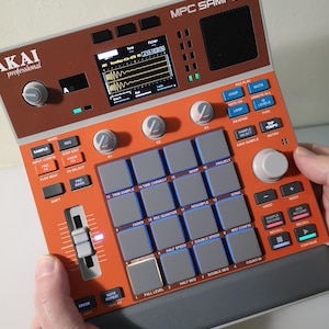 May include: An Akai MPC Sample music production device. The device has an orange and brown colour scheme with numerous buttons, knobs, and a small screen displaying a waveform. The device is held by hands.