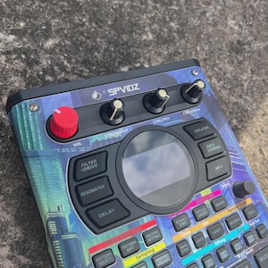 May include: Electronic music production device with a cityscape design. Features knobs, buttons, and a central display. The device has a red knob, and the text "X SPVIDZ" is visible. It is designed for music production and sound manipulation.